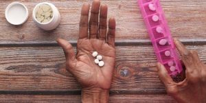 an open hand, palm facing up, laid out on a table. four pills rest in the palm. a pill counter and an open pill bottle flank the hand.