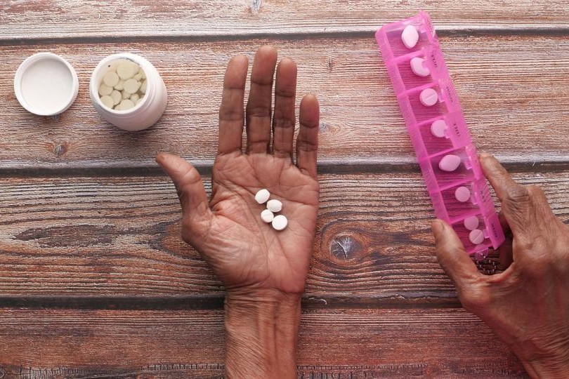 an open hand, palm facing up, laid out on a table. four pills rest in the palm. a pill counter and an open pill bottle flank the hand.