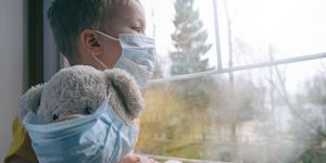 child with teddy bear looks out the window of a suburban home; both child and bear are wearing masks