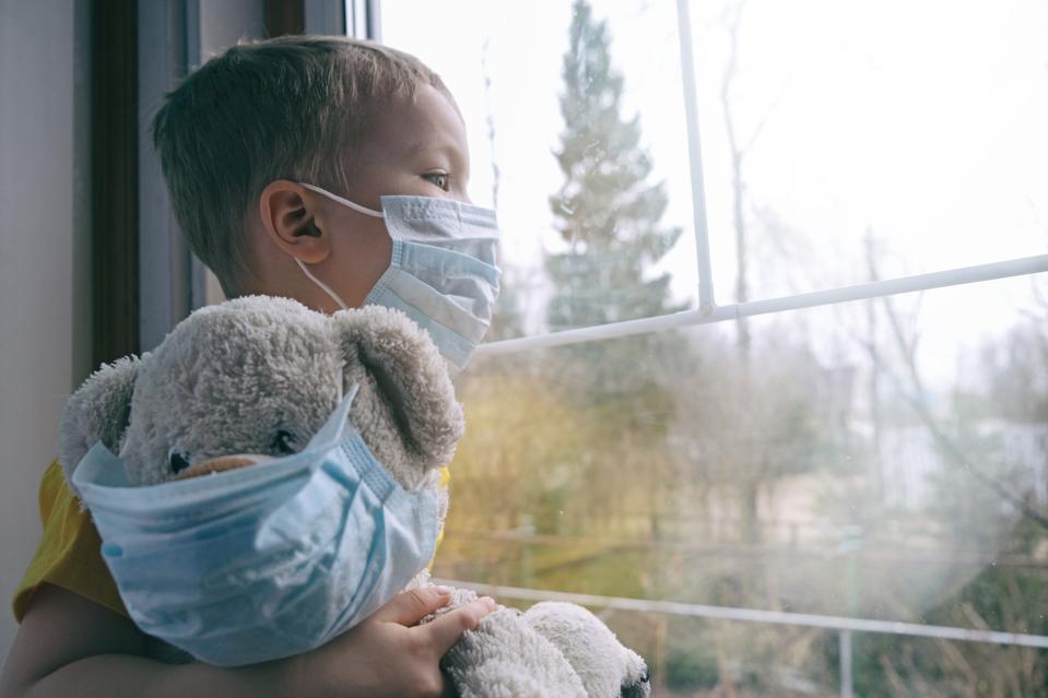 child with teddy bear looks out the window of a suburban home; both child and bear are wearing masks
